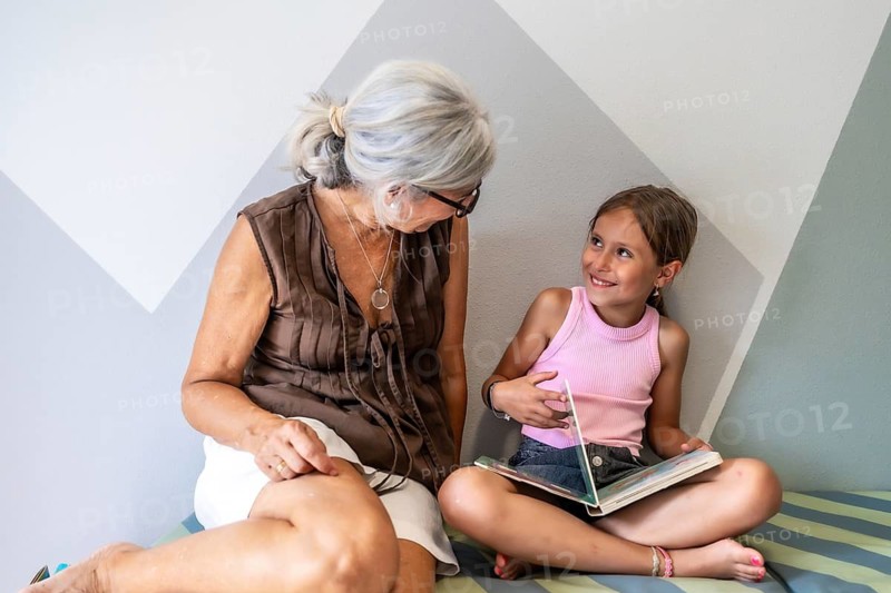 Grandmother and granddaughter are sitting close, sharing a moment of connection while reading a book and smiling at each other, symbolizing intergenerational bonding, childhood, and education