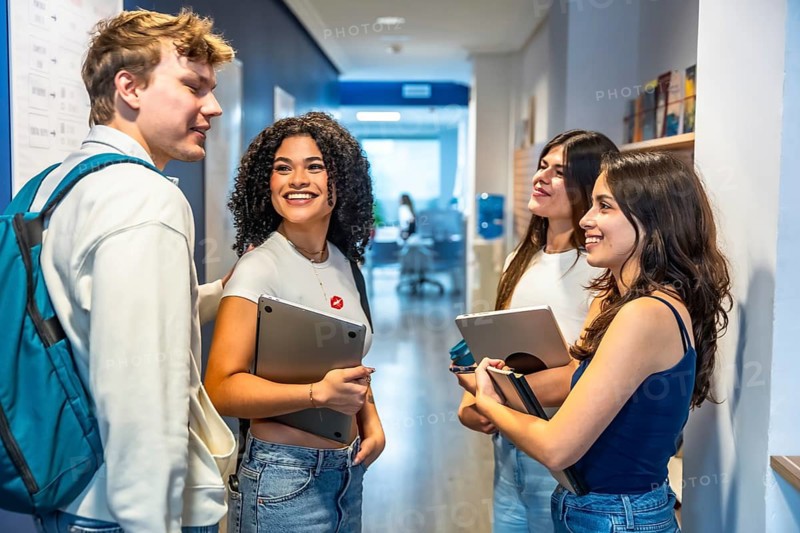Group of multiethnic young university students standing and cheerfully talking in a modern campus hallway, holding laptops and textbooks while engaging in a lively discussion