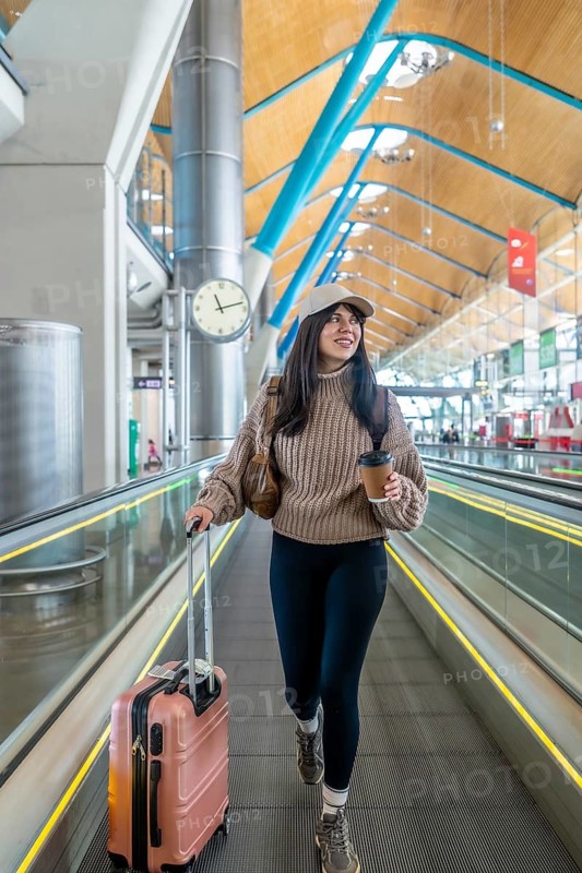 Young woman standing on an automatic moving walkway with a suitcase and a coffee cup, looking happy and anticipating a journey in a modern airport terminal