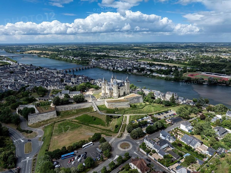 Aerial view of the historic Château de Saumur with its distinctive round corner towers and bright facades, surrounded by well-kept green spaces under a slightly cloudy summer sky, with the town of Saumur on the Loire River, Maine-et-Loire, Pays de la Loire, France