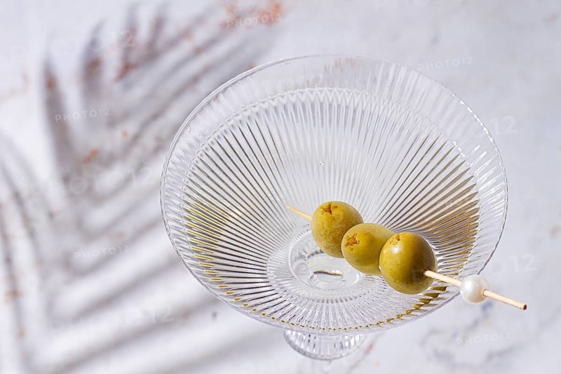 A clear glass cocktail sits on a white marble surface. Three green olives are placed on a skewer resting inside the glass. The light shines on the glass, highlighting its shape
