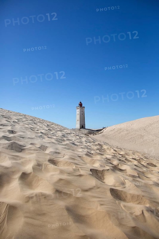 A lighthouse rises amidst wide sand dunes under a bright blue sky, lighthouse in the Rubjerg Knude hiking dune, Jutland, Denmark