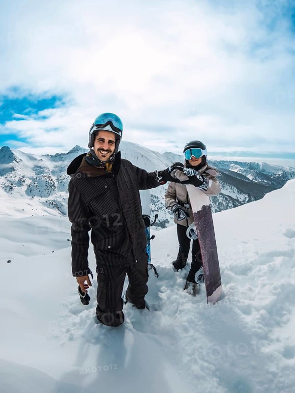 Happy couple on an alpine peak holding snowboards and fist bumping, smiling under a bright blue sky while enjoying a sunny winter snowboarding adventure together