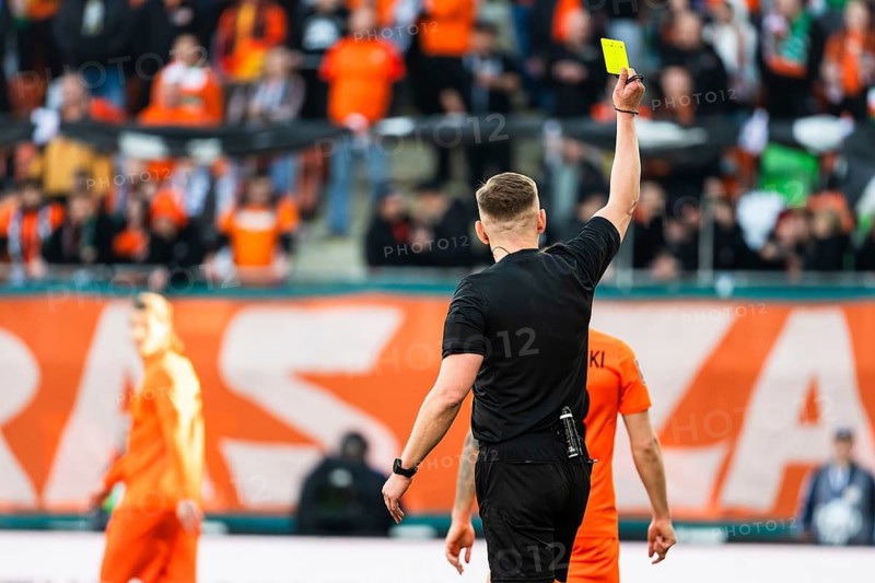 Back view of a soccer referee in a black uniform holding up a yellow card for a foul during a professional match in a crowded stadium in Lubin, Poland