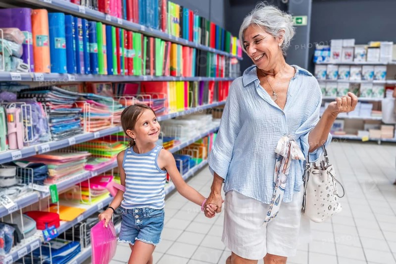 Smiling grandmother and granddaughter holding hands walking through a colorful supermarket aisle, choosing binders and school supplies together for the new academic year