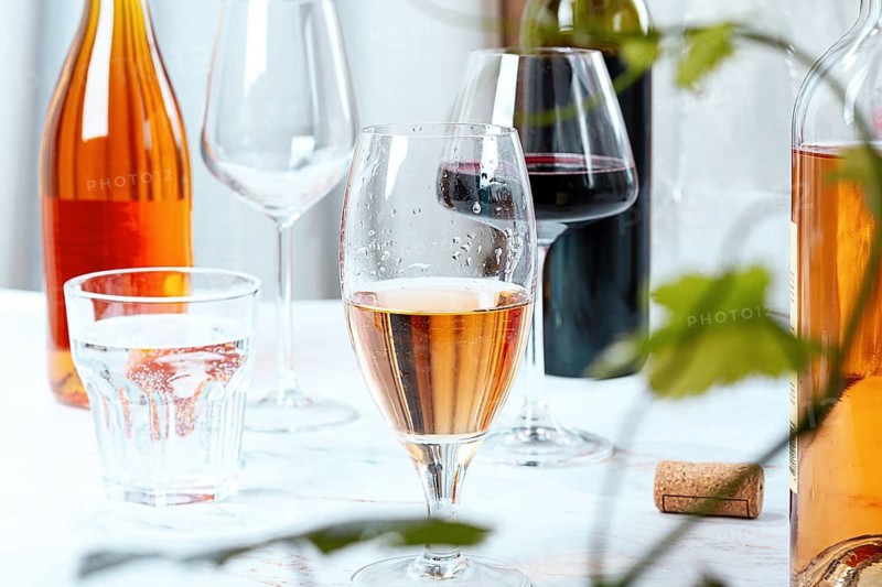 Several glasses and bottles are placed on a table. A glass with rose wine is in focus, next to a glass of water and red wine. The scene is casual and resembles a social event