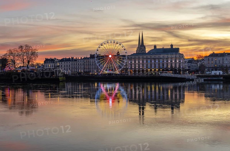 Bayonne, france ferris wheel sparkling with lights reflecting in the adour river water, showcasing the illuminated city buildings and cathedral spires under a colorful sunset sky during christmas