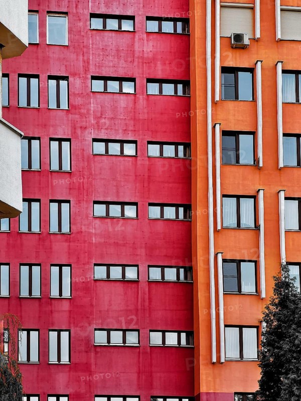 Fragments of modern urban architecture. The red and orange walls of high-rise buildings, Minsk, Belarus. Two modern buildings stand side by side, one with bright red facade and other with vivid orange facade. Rectangular windows form repeating grid across both structures. Orange building features white vertical elements and air conditioning unit near top. The scene highlights bold color contrast and clean urban geometry