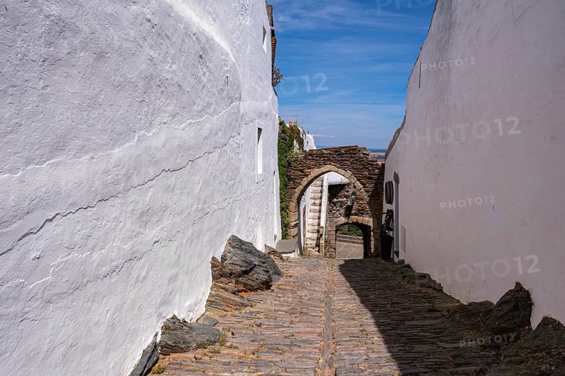 Narrow alley with paved path and white painted walls, leading to a rustic archway, Monsaraz, Évora, Portugal