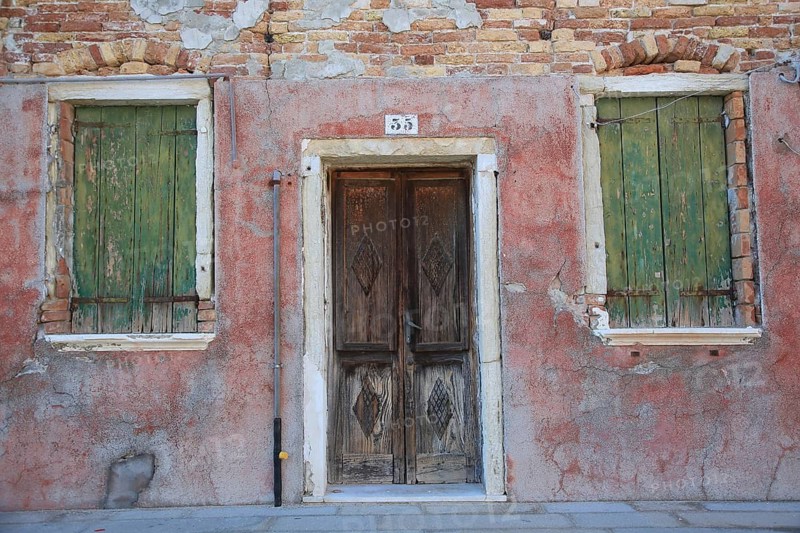 Two windows and a door in an old, weathered red brick façade, windows with green shutters, colorful houses on the island of Burano, Venice, Veneto, Italy