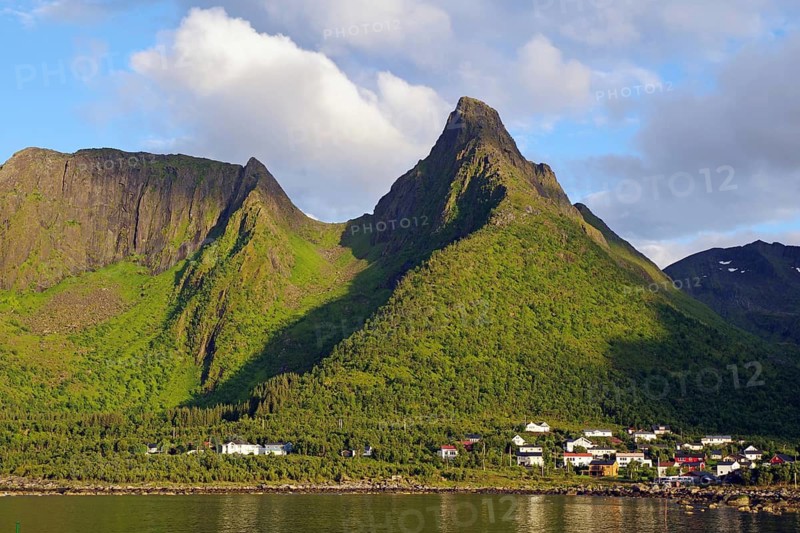 A village at the foot of a green mountain under a cloudy sky. Various houses are visible, Mefjordvaer, Senja, Troms, Norway