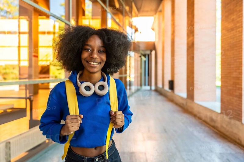 Young black woman with an afro hairstyle, wearing a blue hoodie, yellow backpack, and headphones around her neck, smiling confidently while standing outdoors on a university campus