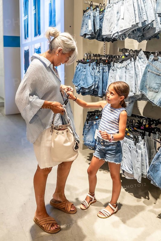 Grandmother and granddaughter bonding while trying on clothes in a bright retail store with racks of denim shorts, enjoying a multi generational shopping experience together