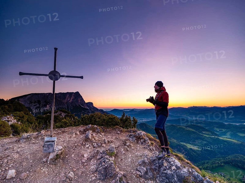 Mountaineer with camera at dawn at the summit cross of Rauhen Kopf, in the back of Untersberg, Berchtesgaden Alps, Bischofswiesen, Berchtesgadener Land, Upper Bavaria, Germany