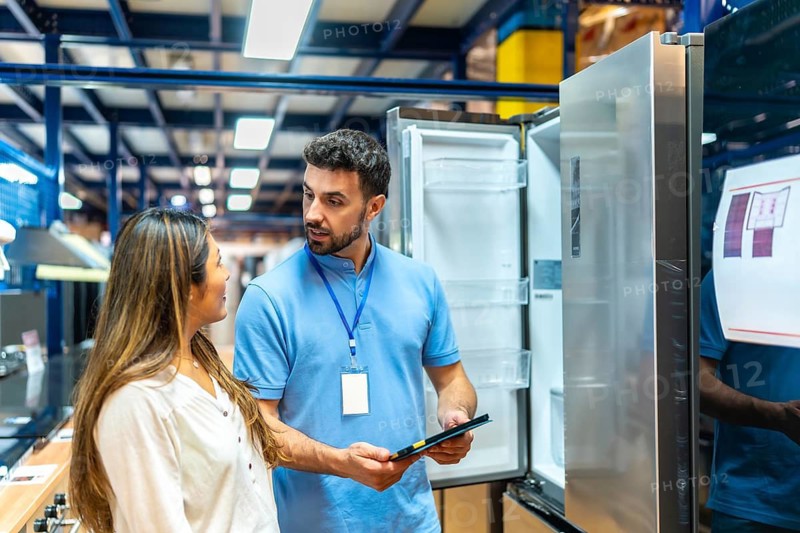 Salesperson showing appliance features to female customer in a retail store, discussing product specifications while shopping for a new refrigerator, symbolizing consumer choices and home upgrade