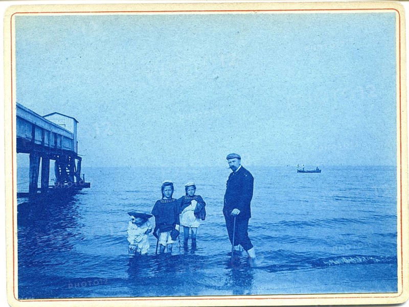 Bathing, swimwear, leisure, summer, cyanotype, man with girl, children, man and children standing by the sea next to a wooden pier with a distant boat, around 1908