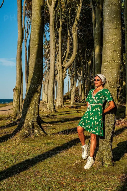 A woman (54) in a green tennis dress and white trainers is leaning casually against a tree, gazing into the distance, Gespensterwald, Nienhagen, Germany