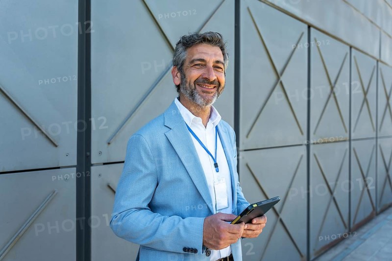 Smiling businessman wearing a blazer and id lanyard looking at camera while holding a digital tablet with both hands, standing in front of a modern building wall