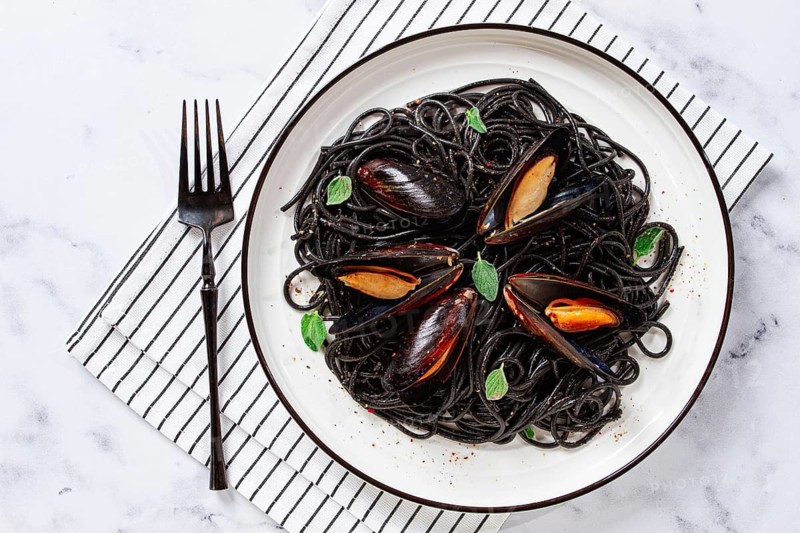 Black spaghetti with mussels, on a white plate with a fork, on a striped napkin, marble background, homemade, no people