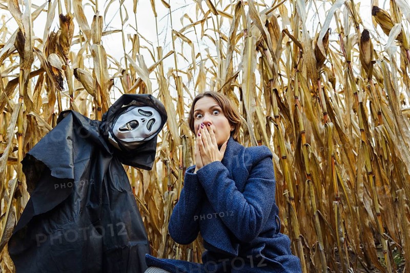 Woman with frightened face near figure with death mask in cornfield on Halloween