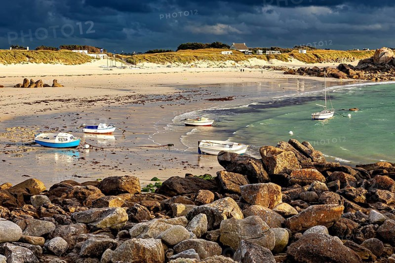 Beach with boats on water and rocks in the foreground under a cloudy sky, The coastal landscape near Cleder in Brittany
