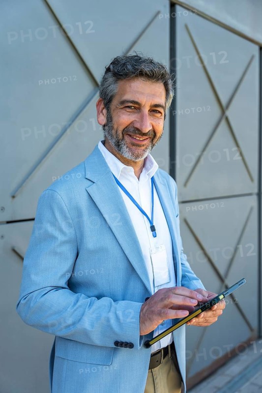 Mature businessman wearing a light blue suit and id badge, holding a digital tablet and smiling, representing professional communication and networking outdoors