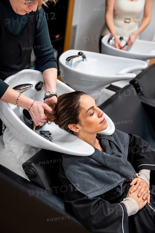 Hairdresser gently washing a relaxed woman's long dark hair at a modern salon sink, providing professional shampoo, head massage and pampering beauty treatment for well being