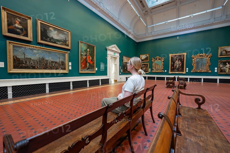 A visitor looks at paintings in a room with green walls, wooden benches and skylight, National Gallery of Ireland, Dublin, Ireland
