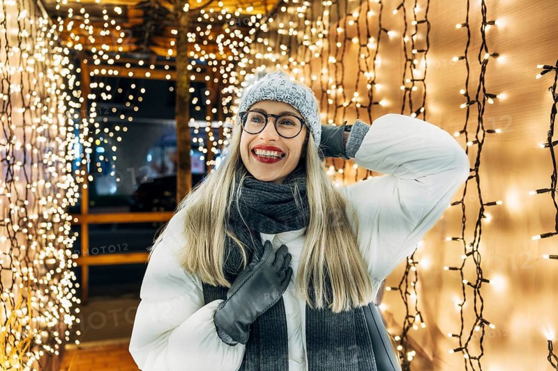 Happy young woman in glasses smiling in winter clothing under festive lights. A woman wearing a gray knit hat, glasses, a white winter coat, dark scarf and gloves stands inside a tunnel of warm white string lights hanging vertically along wooden frames. The dense illumination creates a bright festive glow around her, highlighting winter textures and the cozy holiday Christmas atmosphere of a nighttime seasonal display