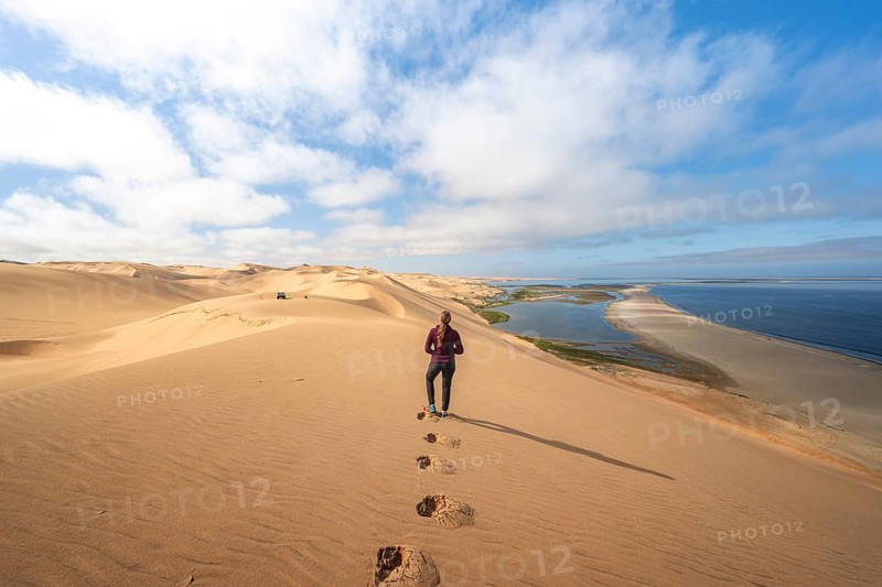 Woman in front of desert landscape, sand dunes and sea, Atlantic coast and sandy desert with sea of dunes, view of lagoon at Sandwich Harbour, Namib Naukluft Park, Namib Desert, Namibia