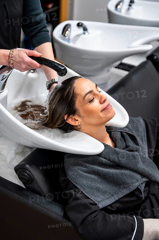 Woman reclining with eyes closed as stylist shampoos and massages her hair at a modern salon sink, enjoying a relaxing, professional hair treatment and pampering session