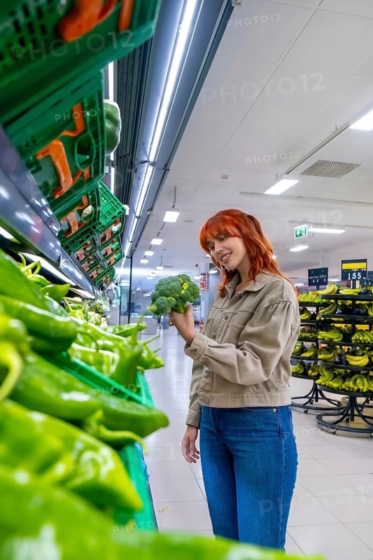 Redhead woman enjoying grocery shopping, selecting healthy green vegetables in the produce section of a modern store, focusing on nutritious food for a balanced diet