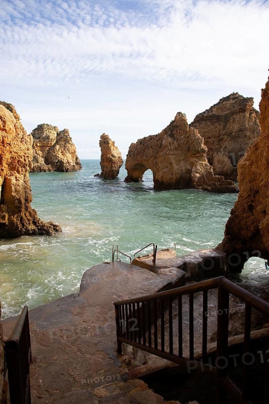 A stone staircase leads down to a rough coastline with rock arches and the blue sea in the background, rocky sandstone coast, rock formations in the sea, Algarve, Lagos, Portugal