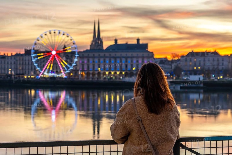 Woman standing on a bridge, overlooking the nive river with the illuminated ferris wheel and city reflections during a vibrant christmas evening sunset in bayonne, france