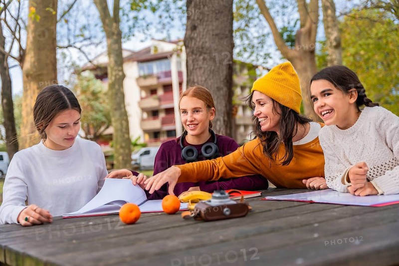 Family consisting of a mother and her three daughters learning and studying together with books and fresh fruit at a wooden picnic table in an autumn park