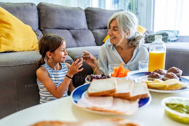 Grandmother and granddaughter joyfully interacting over a meal, sharing muffins and enjoying a casual breakfast or snack time together at home, symbolizing family bonding and affection