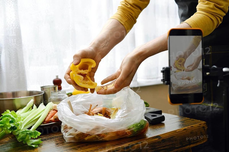 A person peels bell pepper from seeds and throws the waste into a compost bag. Food blogger records an instructional video on his smartphone and explains the importance of using leftovers wisely