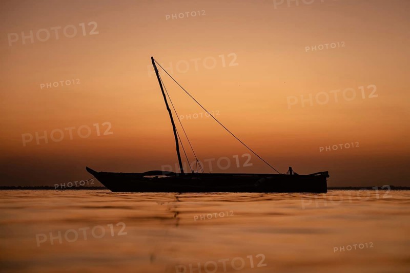 A traditional wooden dhow sailing boat is silhouetted against a calm sea and a deep orange sky during sunset in Zanzibar