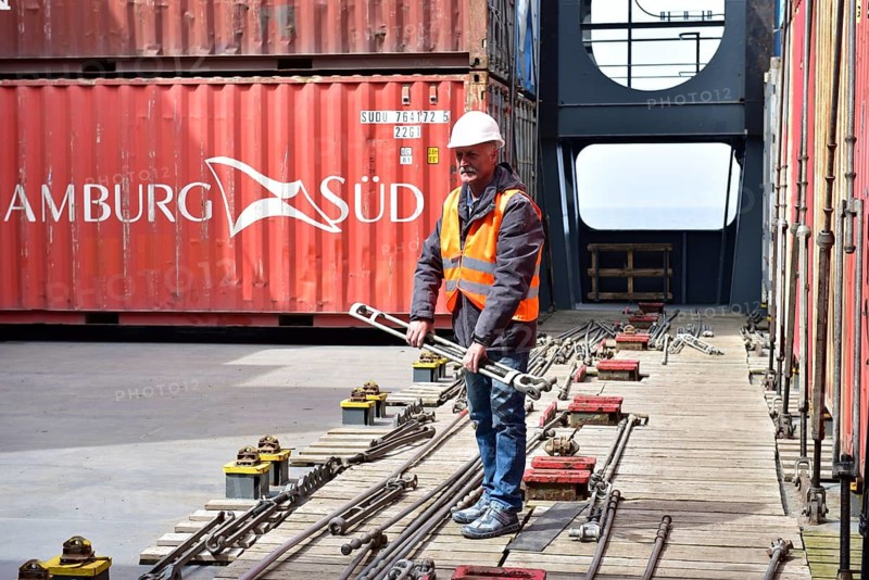 Worker wearing helmet and safety vest holding a large wrench between containers in port, worker holding tie rods on a container ship, Schleswig-Holstein, Germany