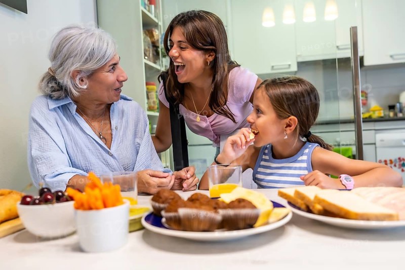 Family members, a grandmother, mother, and granddaughter, bonding and laughing together while sharing a healthy snack and drinks around a kitchen table at home