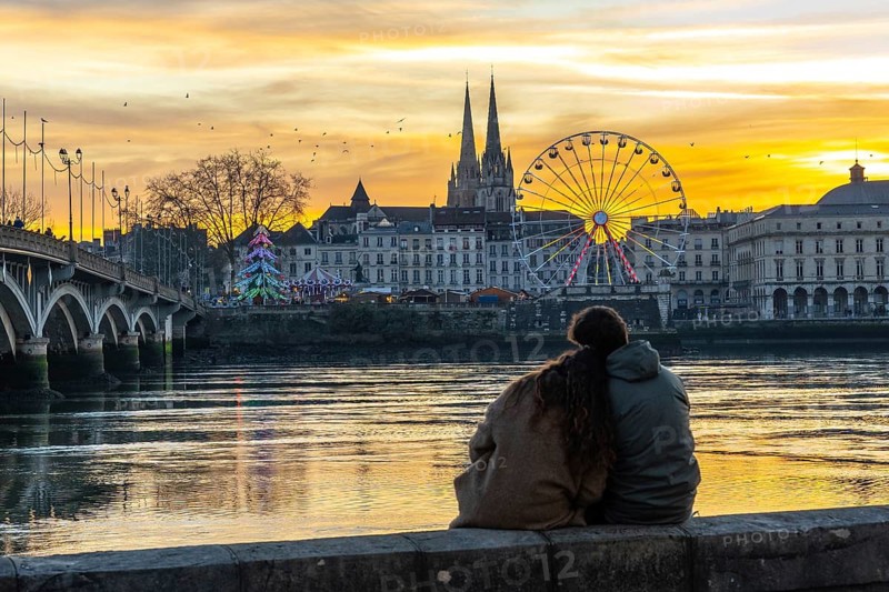 Young couple sits on a wall enjoying a colorful sunset over the adour river in bayonne, france, with ferris wheel, cathedral and illuminated christmas tree reflecting in the water