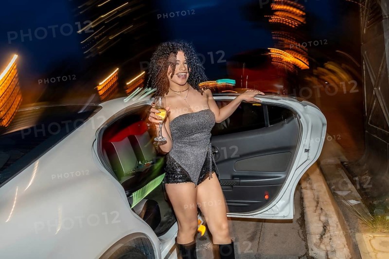 Young black woman stepping out of a premium white car, celebrating with a glass of champagne, embracing the excitement and glamour of a vibrant nightlife event