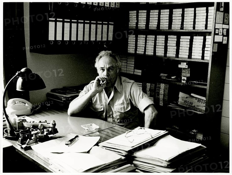 A man sits deep in thought at a desk in a tidy office full of documents and files: Herbert Jacobi (1915–1978), Self-Portrait, 1978