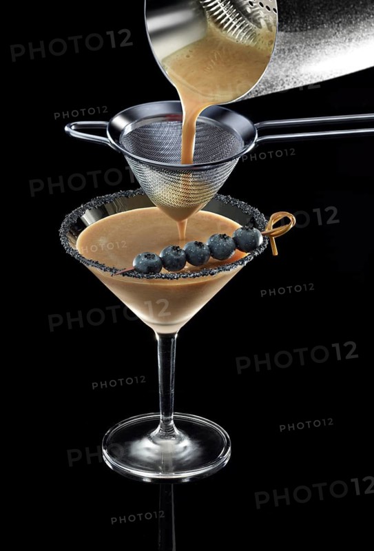 Brown cocktail is poured through a sieve, against a black background, blueberries and black salt as a garnish, studio shot