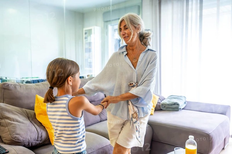 Grandmother and granddaughter smiling and holding hands, sharing a loving moment together in a bright modern living room, representing family bonds and intergenerational happiness