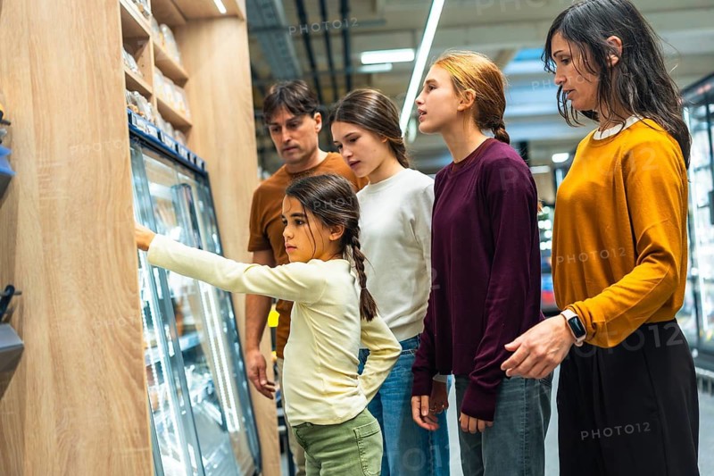 Family group standing in a supermarket aisle, choosing products from a refrigerated display, with a young girl pointing at an item while her parents and sisters observe