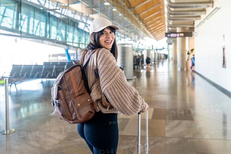 Young woman traveling with backpack and luggage, happily looking back while walking through a bright, spacious airport terminal, anticipating a journey or vacation