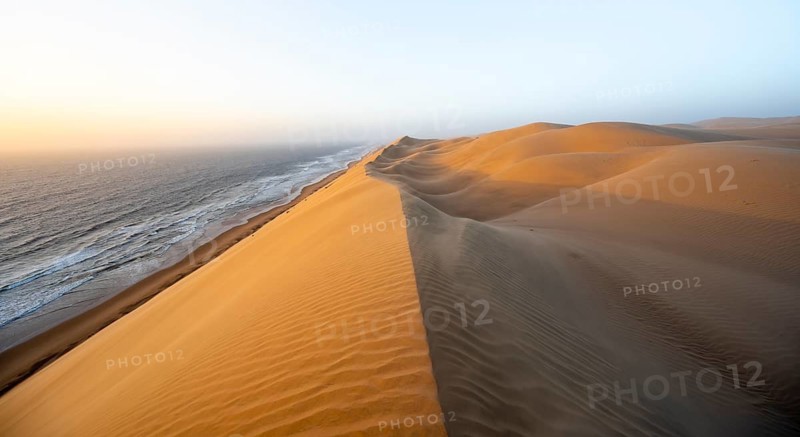 Sand dunes with wave patterns and sea in evening light, wind blowing sand across the dunes, Atlantic coast and sandy desert with sea of dunes, impressive desert landscape at sunset, Sandwich Harbour, Namib Naukluft Park, Namib Desert, Namibia