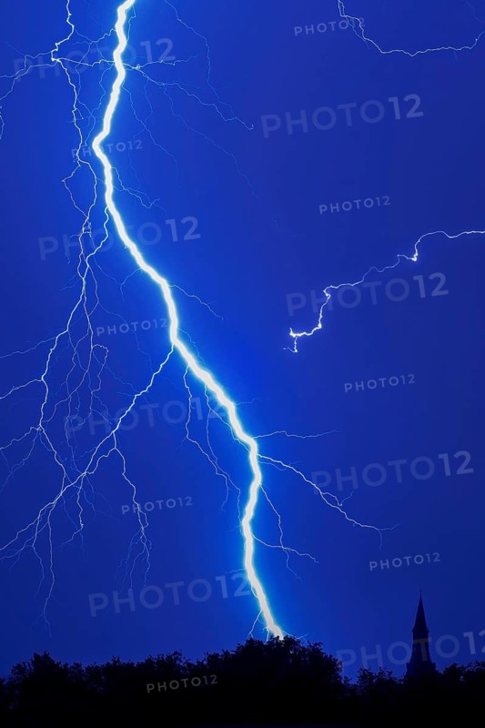 Forked lightning bolt, cloud-to-ground lightning strike during thunderstorm in blue night sky over forest with silhouetted treetops and church tower