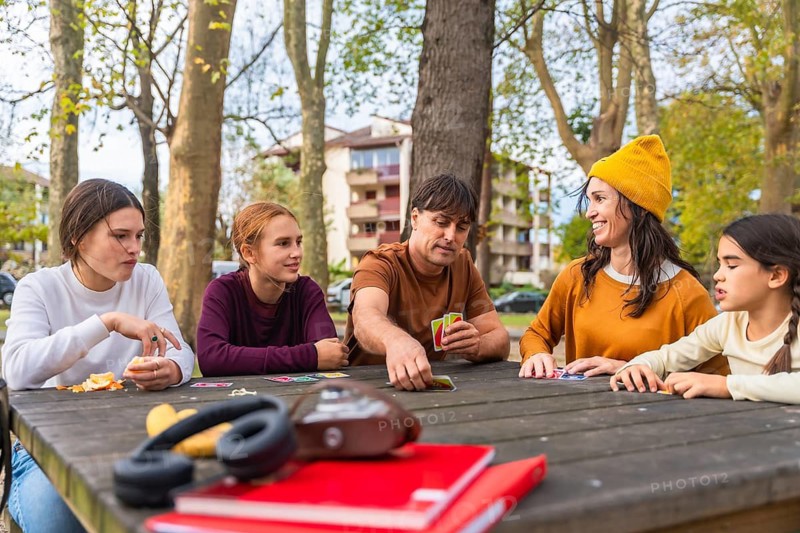 Family playing cards at a picnic table in a park on an autumn day, parents and children laughing and bonding over a casual outdoor game, enjoying quality time together